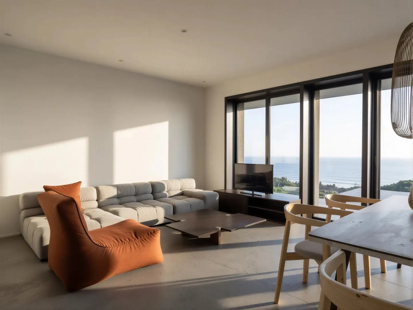 Bright living room with a gray sectional sofa, orange beanbag chair, and a dark wood coffee table. Large windows overlook the ocean.