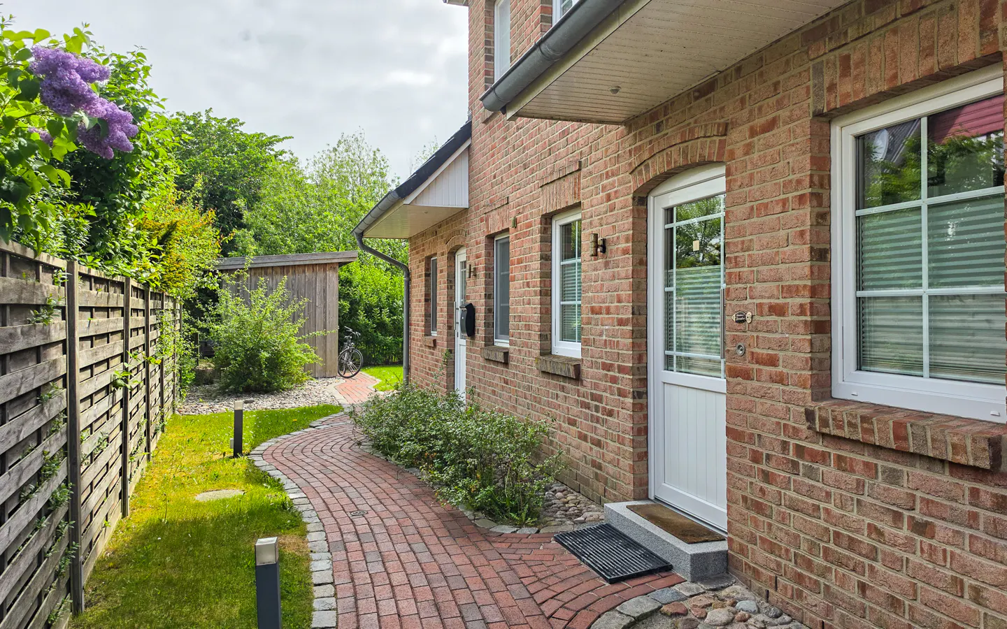 Brick house exterior with a red brick walkway leading to a white front door and a wooden fence with purple flowers.