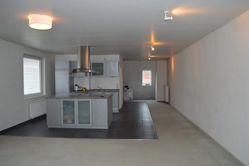 Bright, empty apartment with white walls and a gray kitchen. A stainless steel range hood is above the island. Dark tile separates the kitchen from the living space.