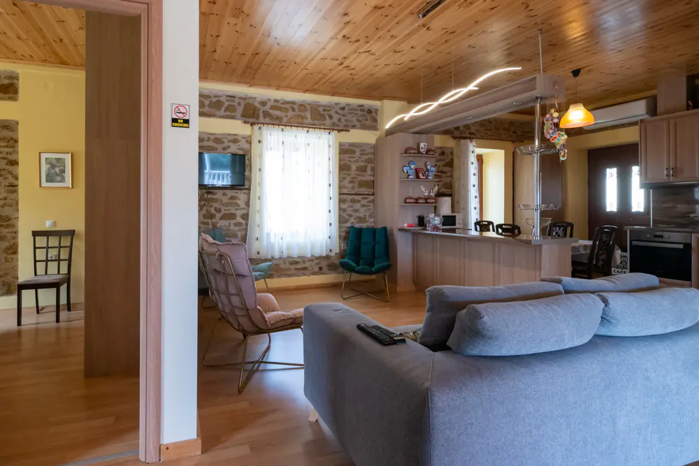 Living room with stone walls, wood ceiling, and gray sofa. A kitchen area is visible in the background.