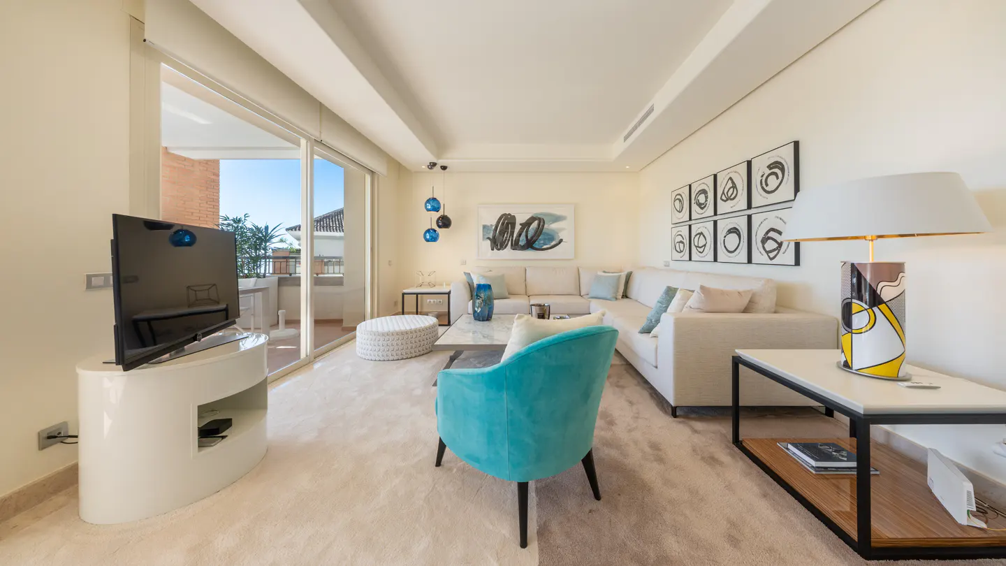 Bright living room with a white sofa, turquoise chair, and a TV on a white stand. A balcony is visible through sliding glass doors.