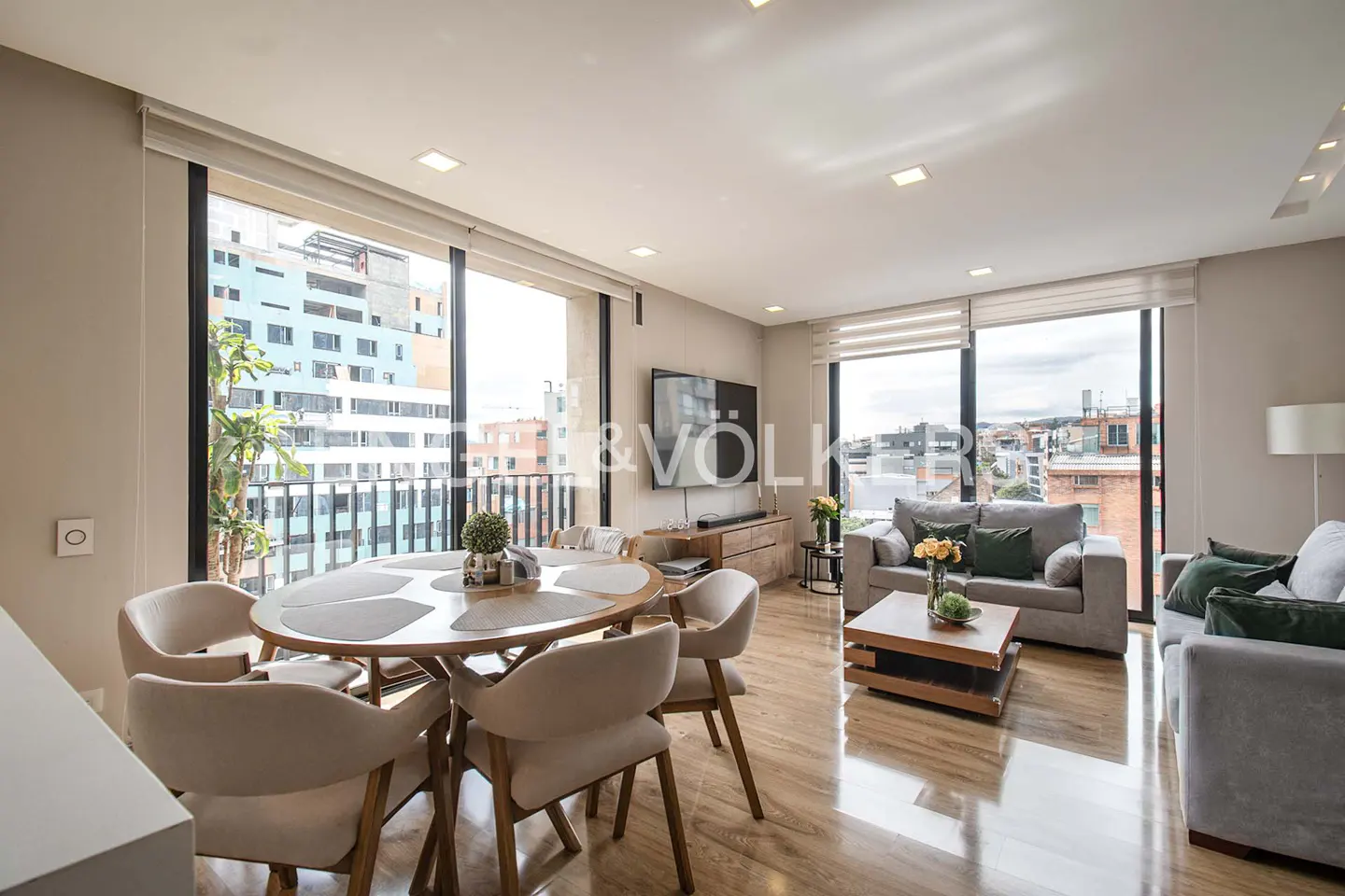 Bright, modern living room with wood floors, a round dining table with six chairs, and two gray sofas. Large windows offer city views.