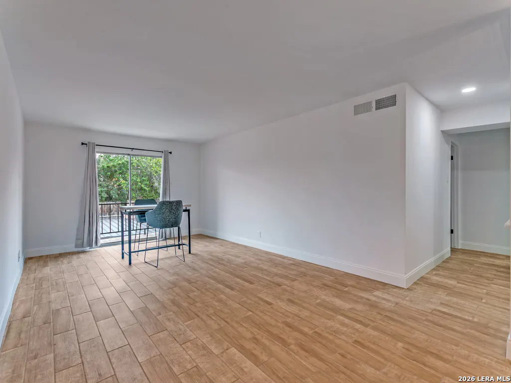 Bright, empty room with wood-look tile flooring, white walls, and a table with two blue chairs by a sliding glass door.