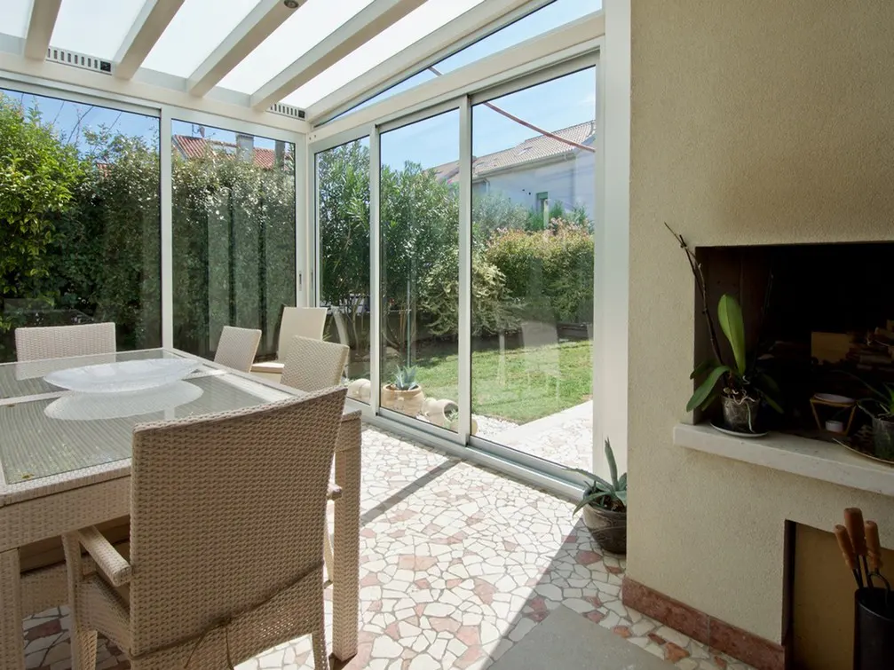 Sunroom with a wicker table and chairs, a fireplace, and a view of the garden through large glass windows.