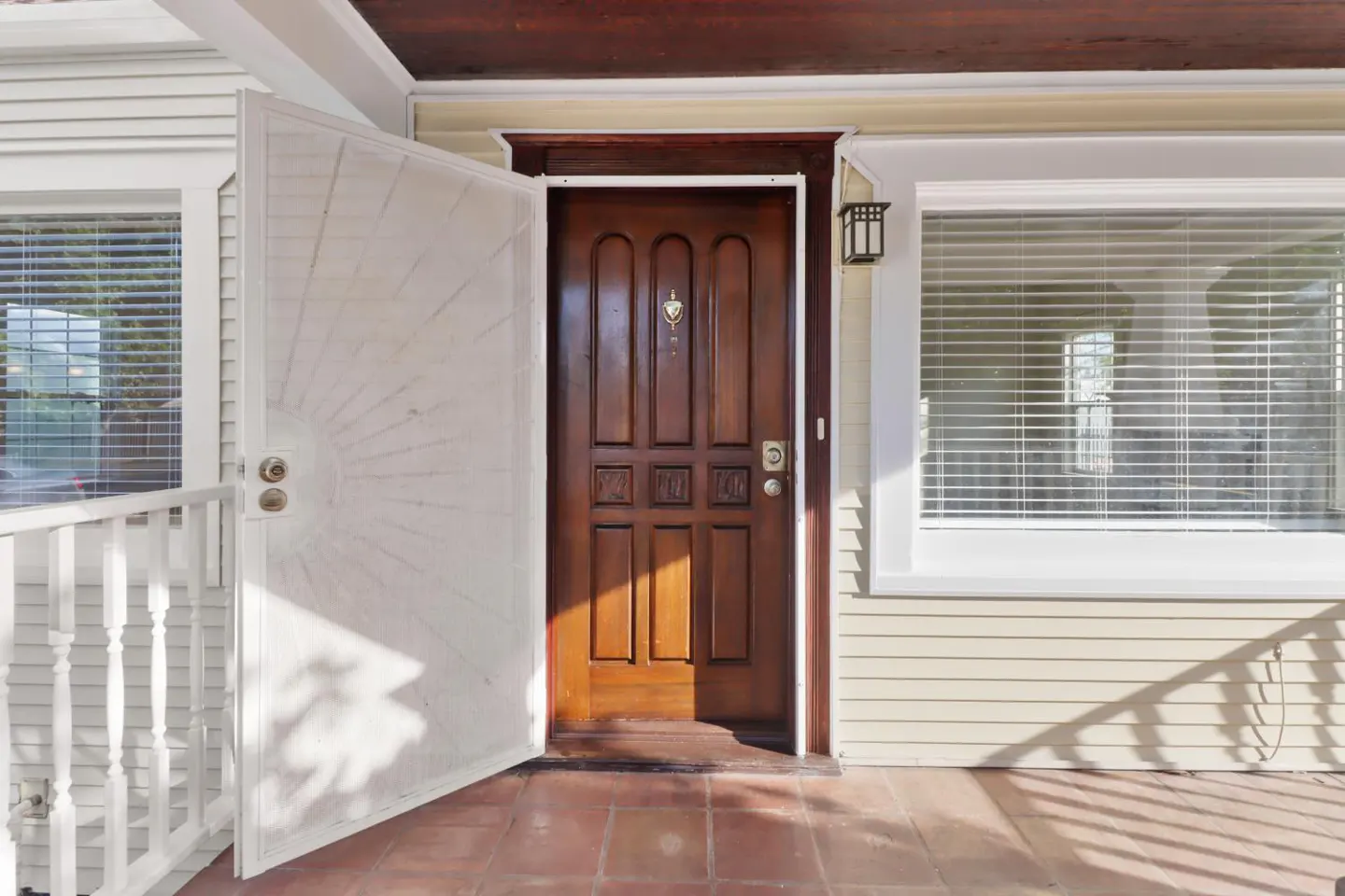 Open brown front door with a white screen door ajar, showing a glimpse of the interior. Windows with blinds flank the door. The porch has red tiles.