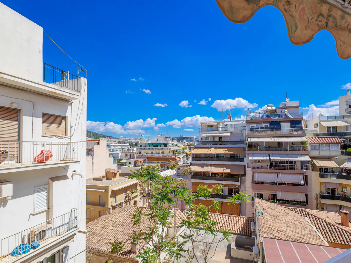 View from a balcony overlooking a city with white buildings, red tile roofs, and a bright blue sky with scattered clouds.