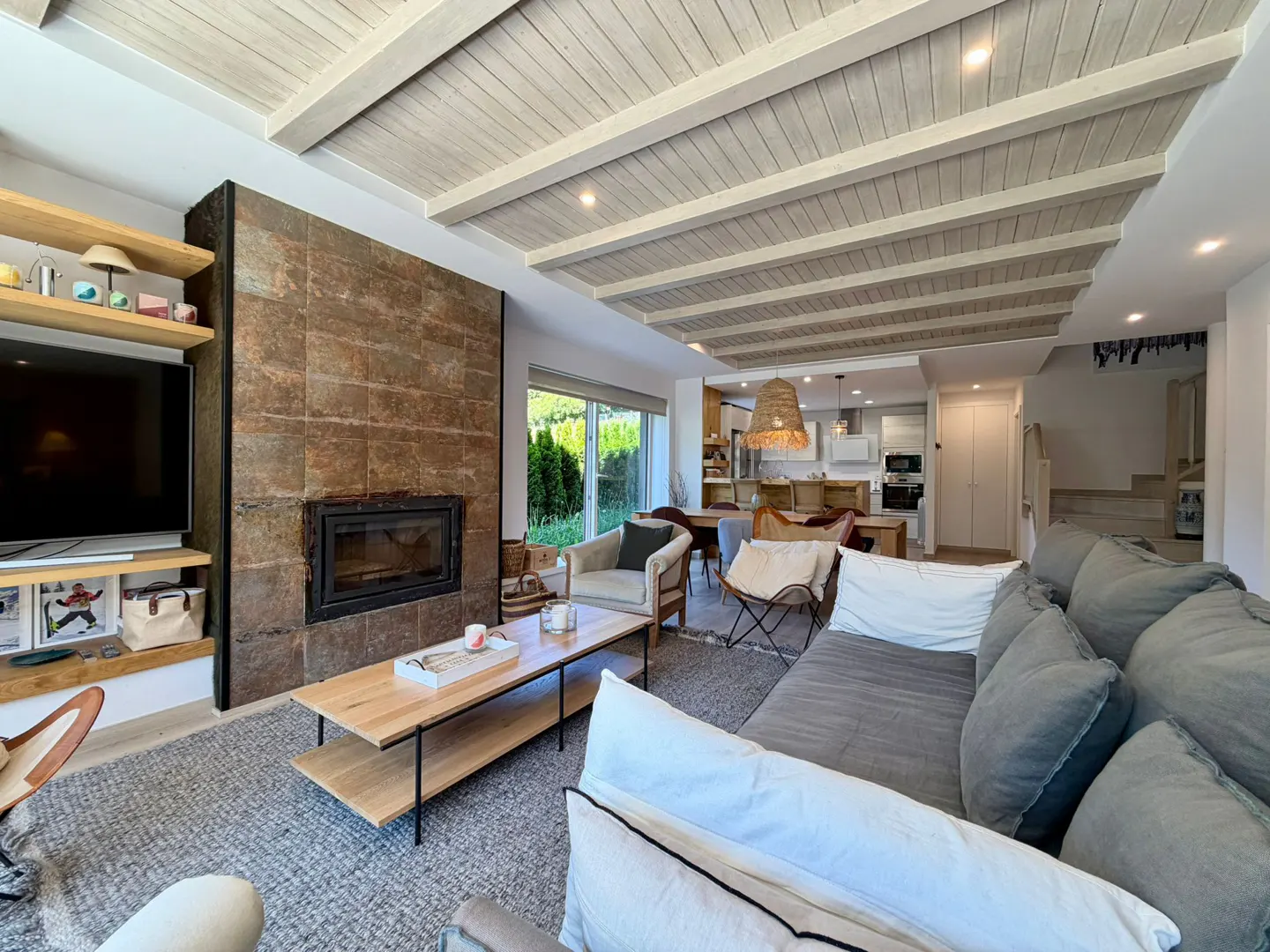 Modern living room with a stone fireplace, gray sofa, wooden coffee table, and exposed beam ceiling leading to a kitchen.