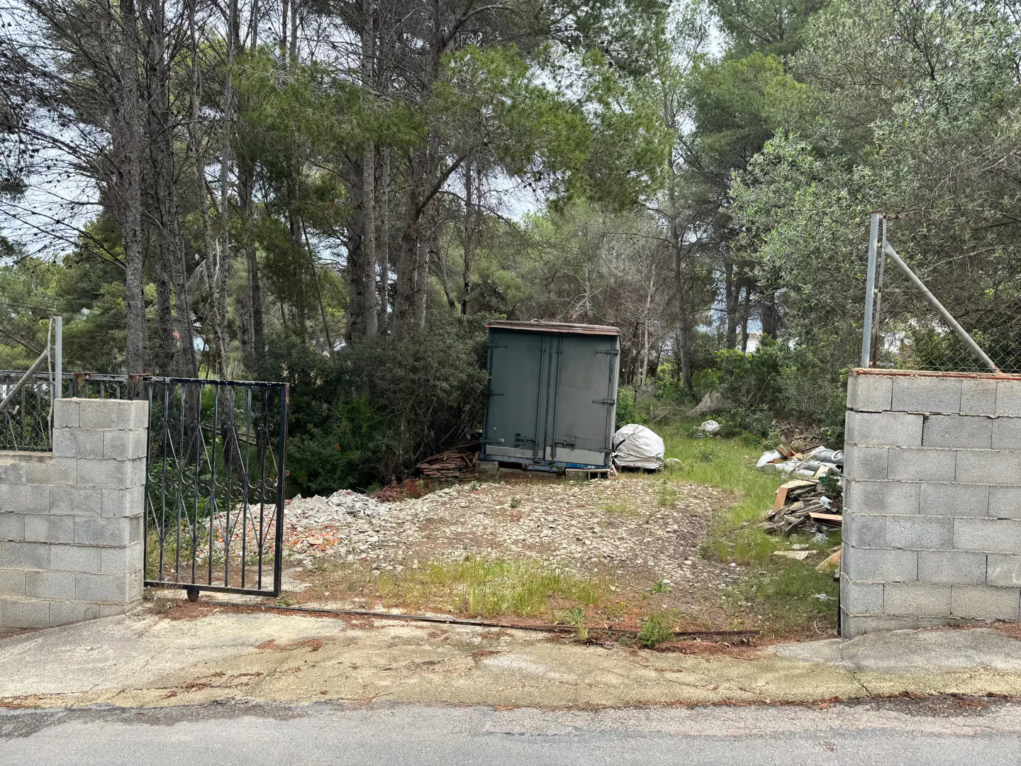 Gated lot with trees in the background. A green container sits on the lot, with debris scattered around.