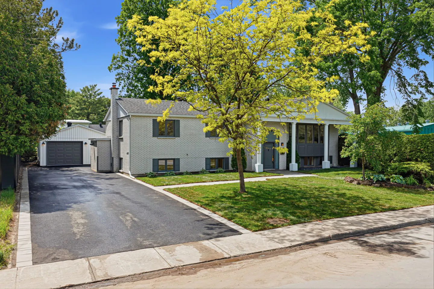 Single-story light gray brick house with a paved driveway, attached garage, and a large tree in the front yard.