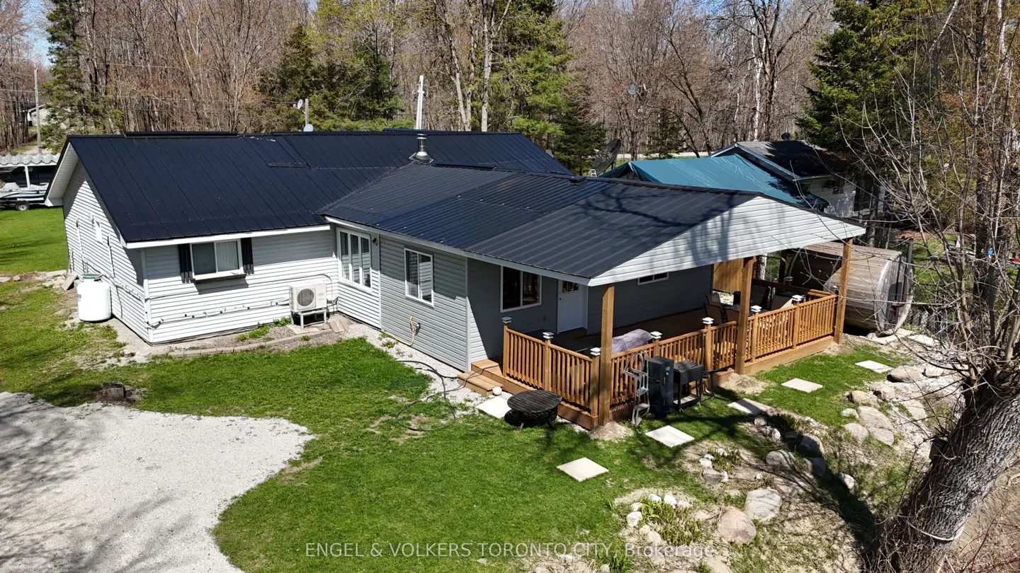 Aerial view of a gray house with a black metal roof, a wooden porch, and a green lawn surrounded by trees.