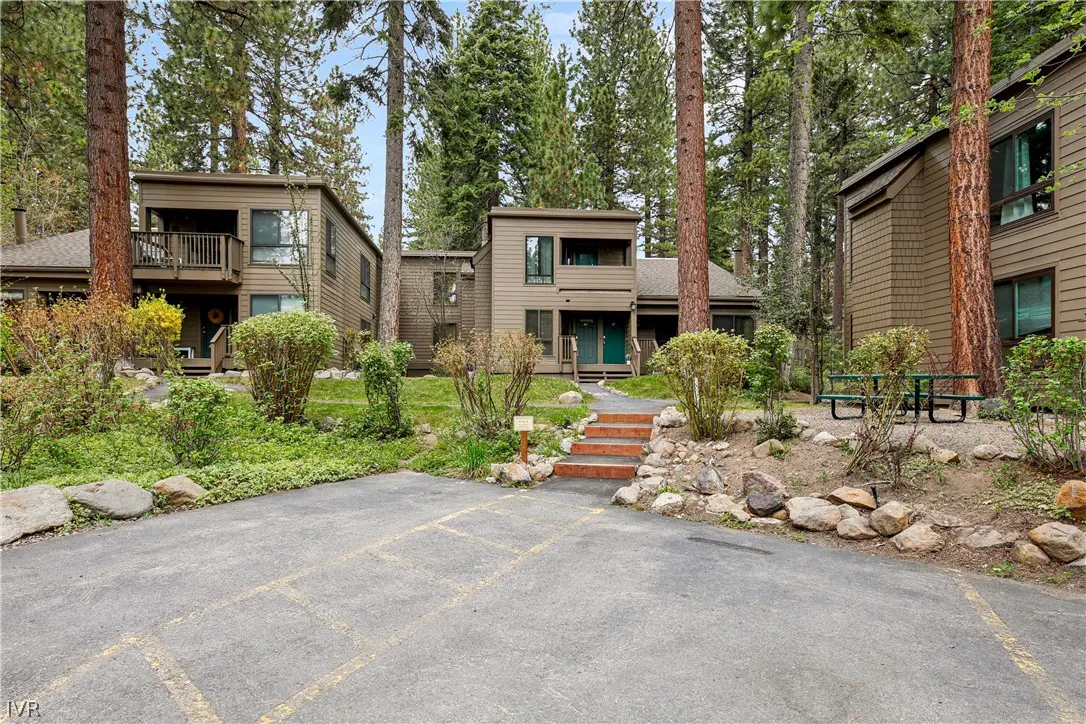 Exterior view of brown two-story townhouses surrounded by tall pine trees and greenery. A parking lot is in the foreground.