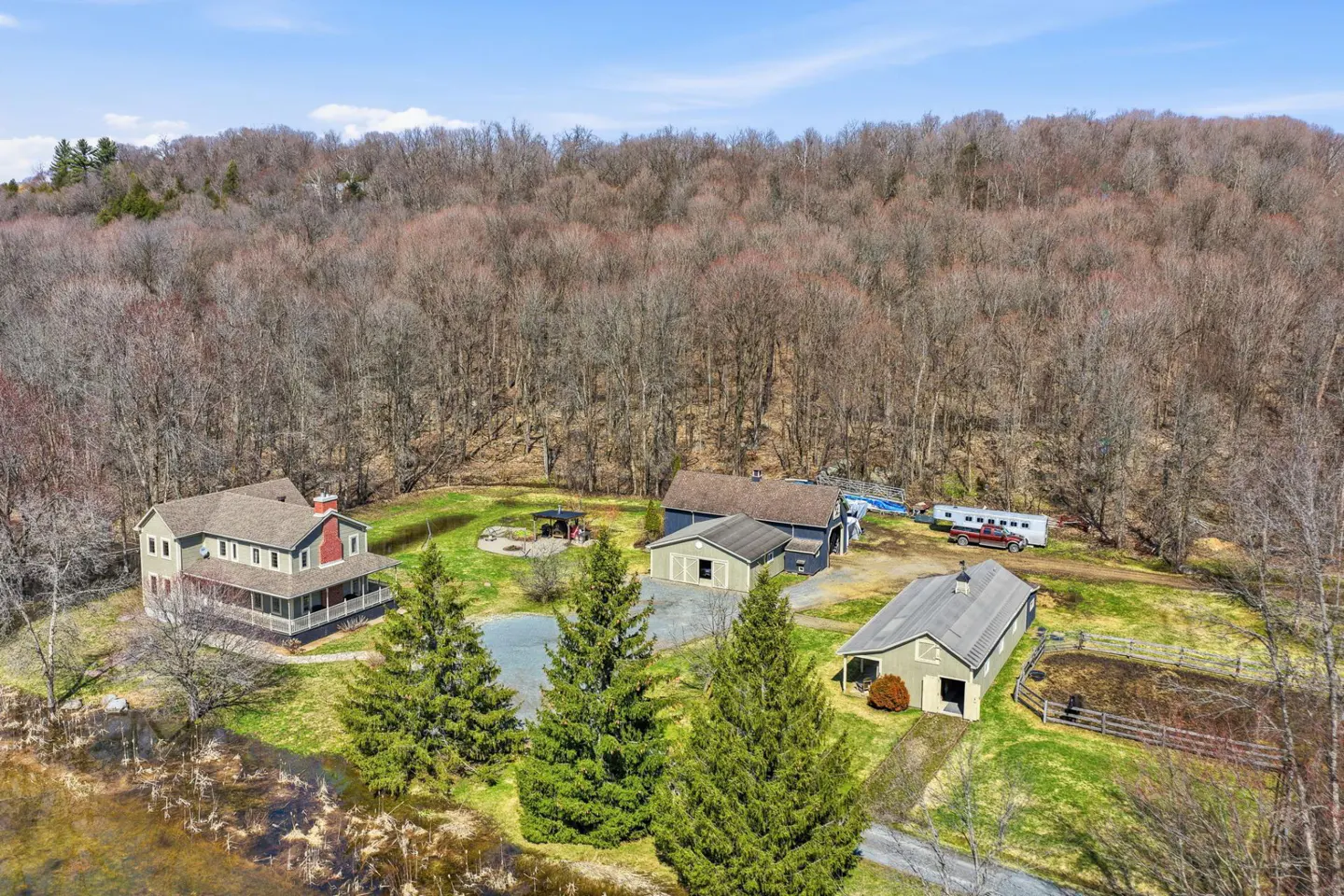 Aerial view of a large property with a house, barns, pond, and wooded area under a blue sky.