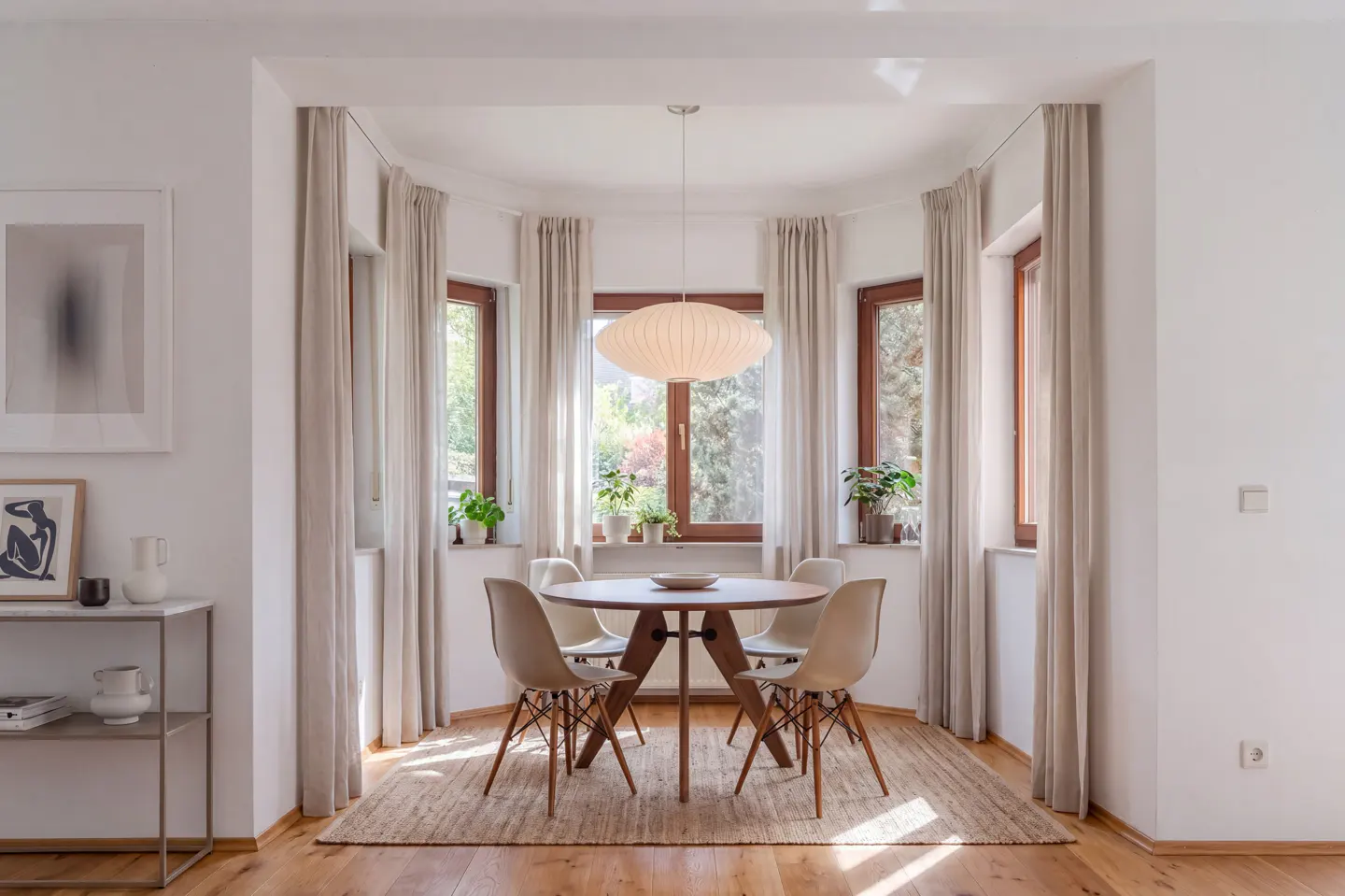 Bright dining room with wood floors, a round table with four chairs, and a modern light fixture. Windows with beige curtains surround the table.