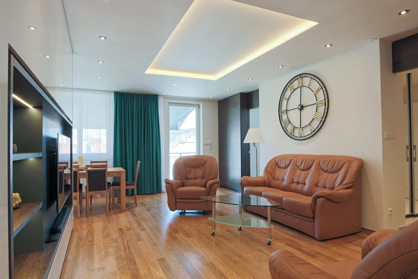 Bright living room with brown leather sofa and chairs, glass table, and large clock on the wall. Teal curtains and wood floors add warmth.