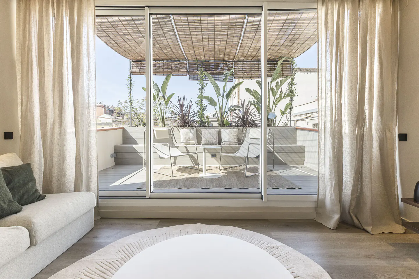 Living room view to a sunny terrace with plants, chairs, and a bamboo shade. Beige curtains frame the sliding glass doors.