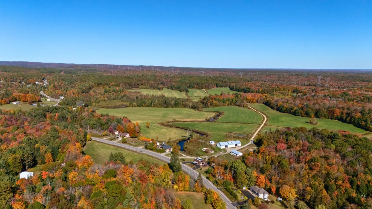 Aerial view of a rural landscape with colorful fall foliage, green fields, and scattered houses under a clear blue sky.