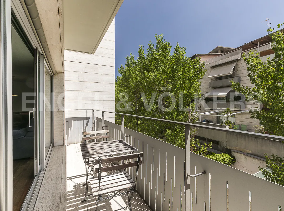 A sunny balcony with a small wooden table and two chairs, overlooking green trees and a neighboring apartment building.