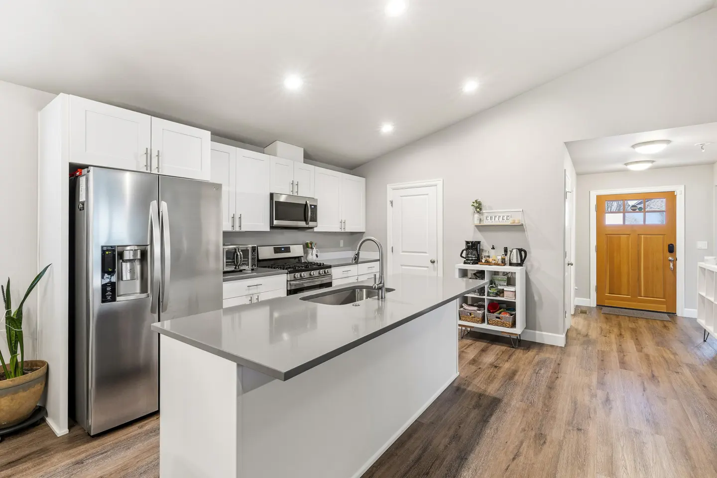 Bright, modern kitchen with white cabinets, stainless steel appliances, and a gray island. Wood-look flooring leads to a wooden front door.