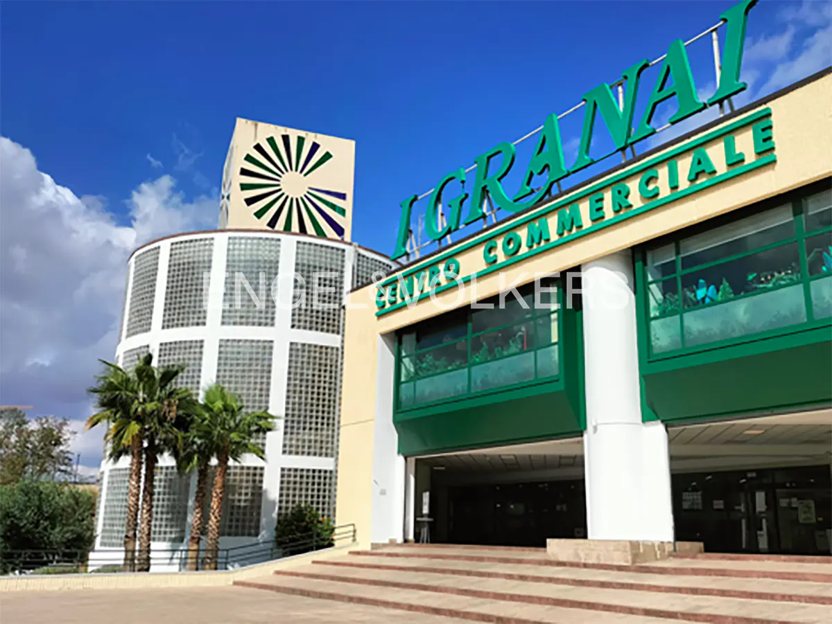 Exterior view of "I Granai" shopping center with green signage, glass block tower, palm trees, and blue sky.