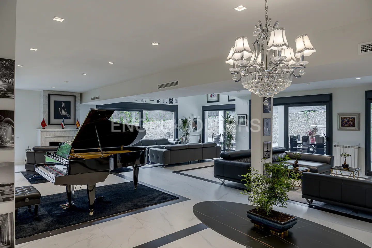 Bright living room with a black grand piano, black sofas, and a crystal chandelier. Large windows offer outdoor views.