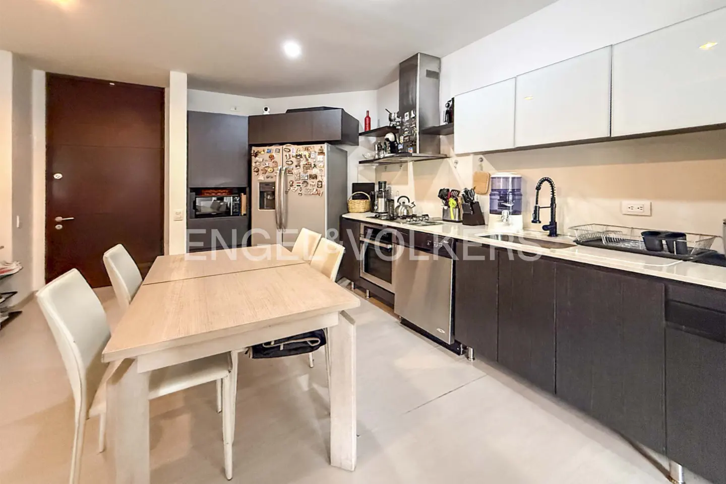 A modern kitchen with white cabinets, dark wood lower cabinets, stainless steel appliances, and a light wood dining table with white chairs.