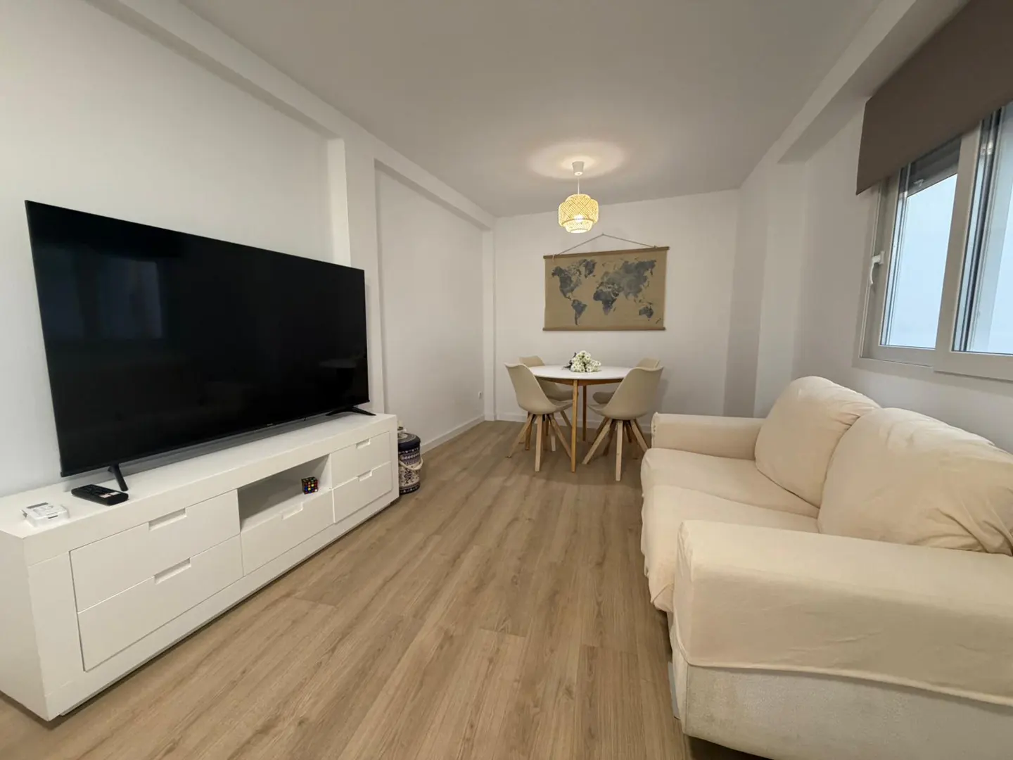 Living room with a large TV on a white cabinet, a beige sofa, and a dining table with four chairs. A world map hangs on the wall.