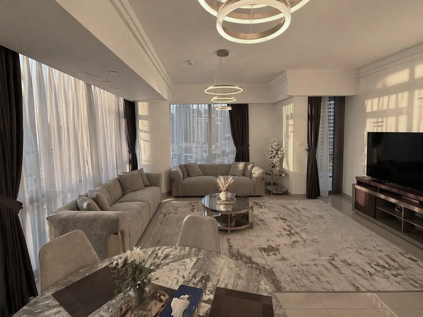 A bright, modern living room with beige sofas, a round marble table, and floor-to-ceiling windows with sheer curtains.