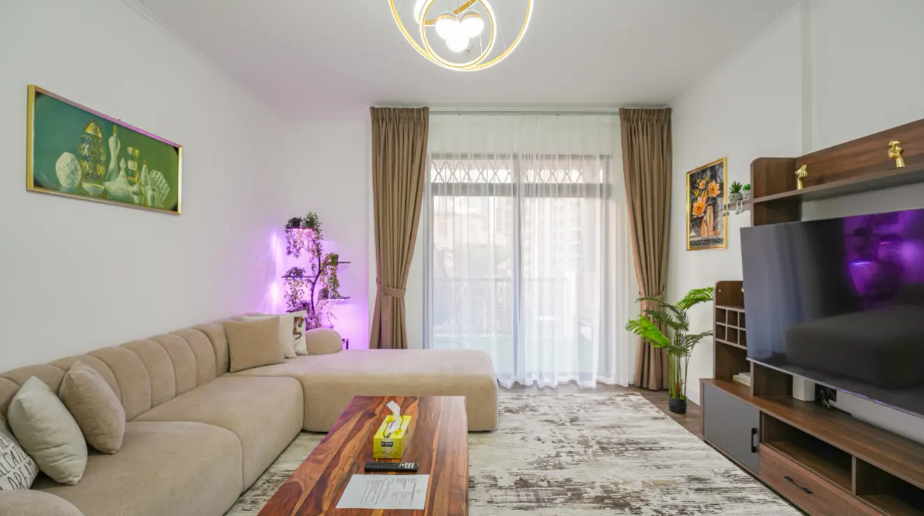 Living room with beige sectional sofa, wood coffee table, and TV stand. Neutral rug and curtains. Artwork on walls, modern light fixture.