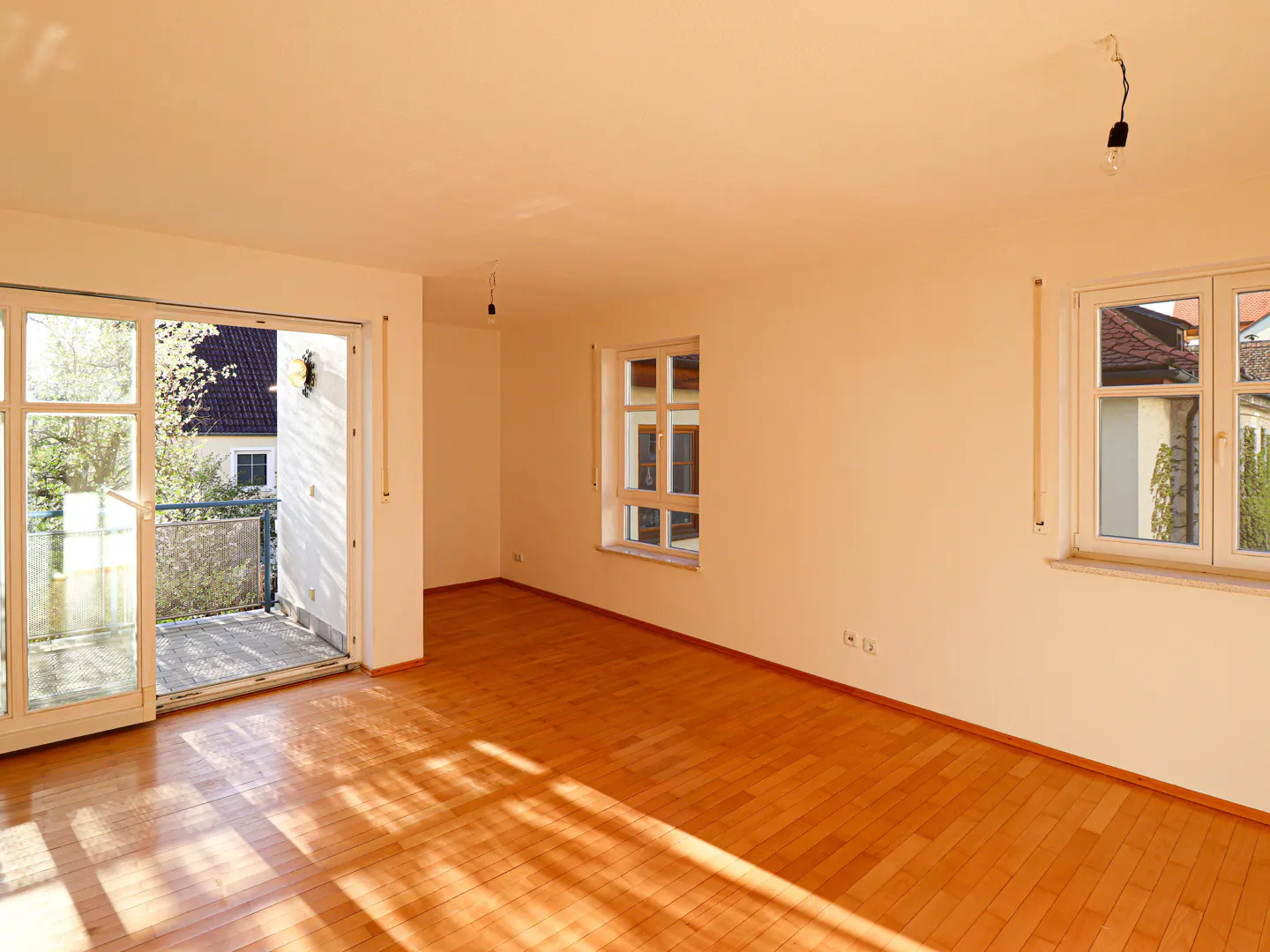 Bright, empty room with hardwood floors, white walls, and natural light. Sliding glass doors open to a balcony. Two windows offer outside views.