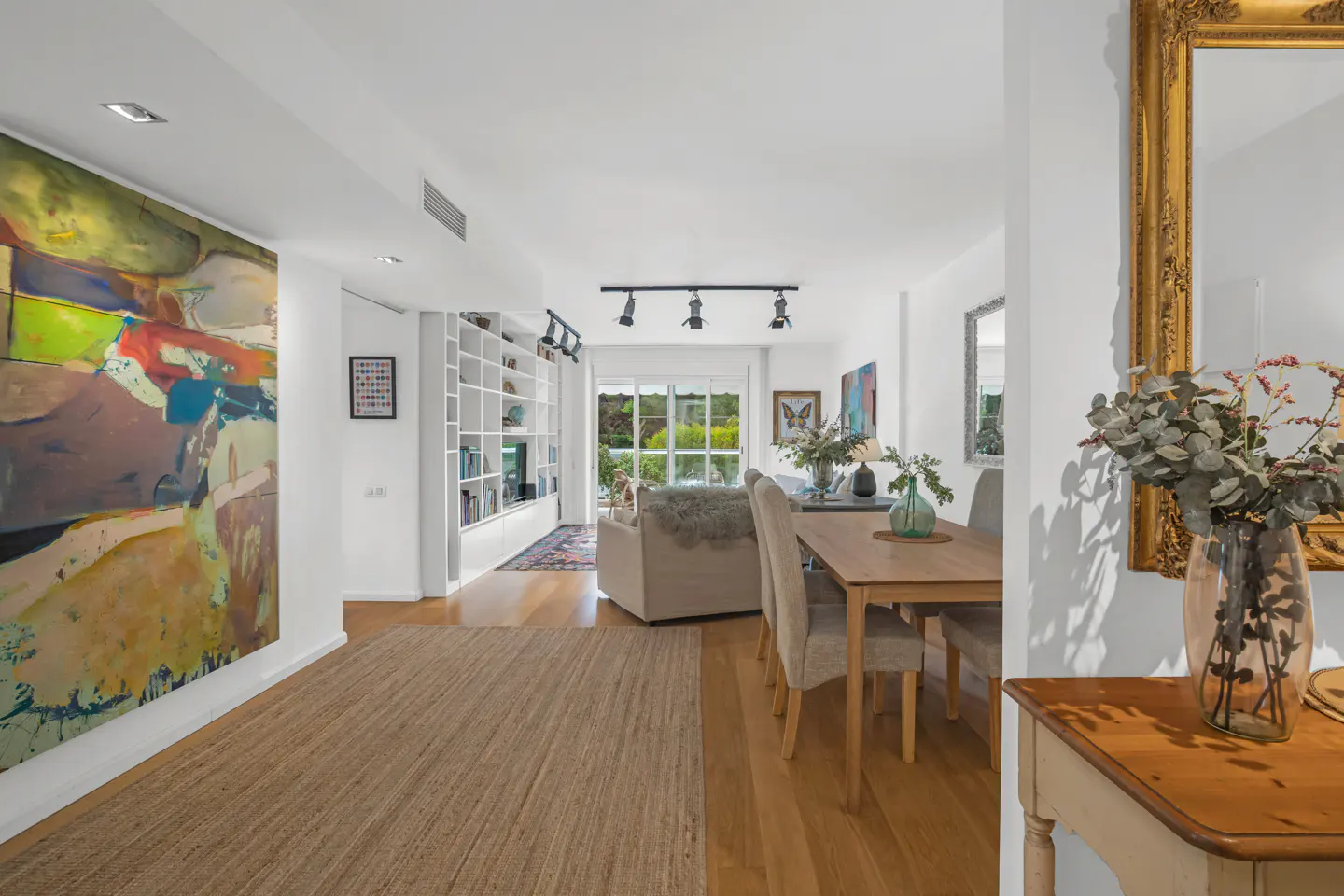 Bright, modern living space with a large abstract painting, dining table, and built-in bookshelves. Natural light fills the room.