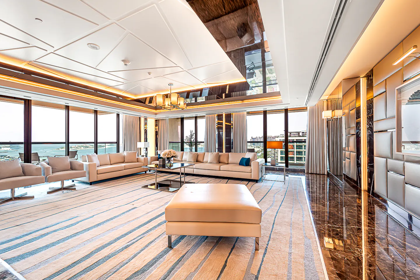 A luxurious living room with beige sofas, a patterned rug, and floor-to-ceiling windows with a city view.