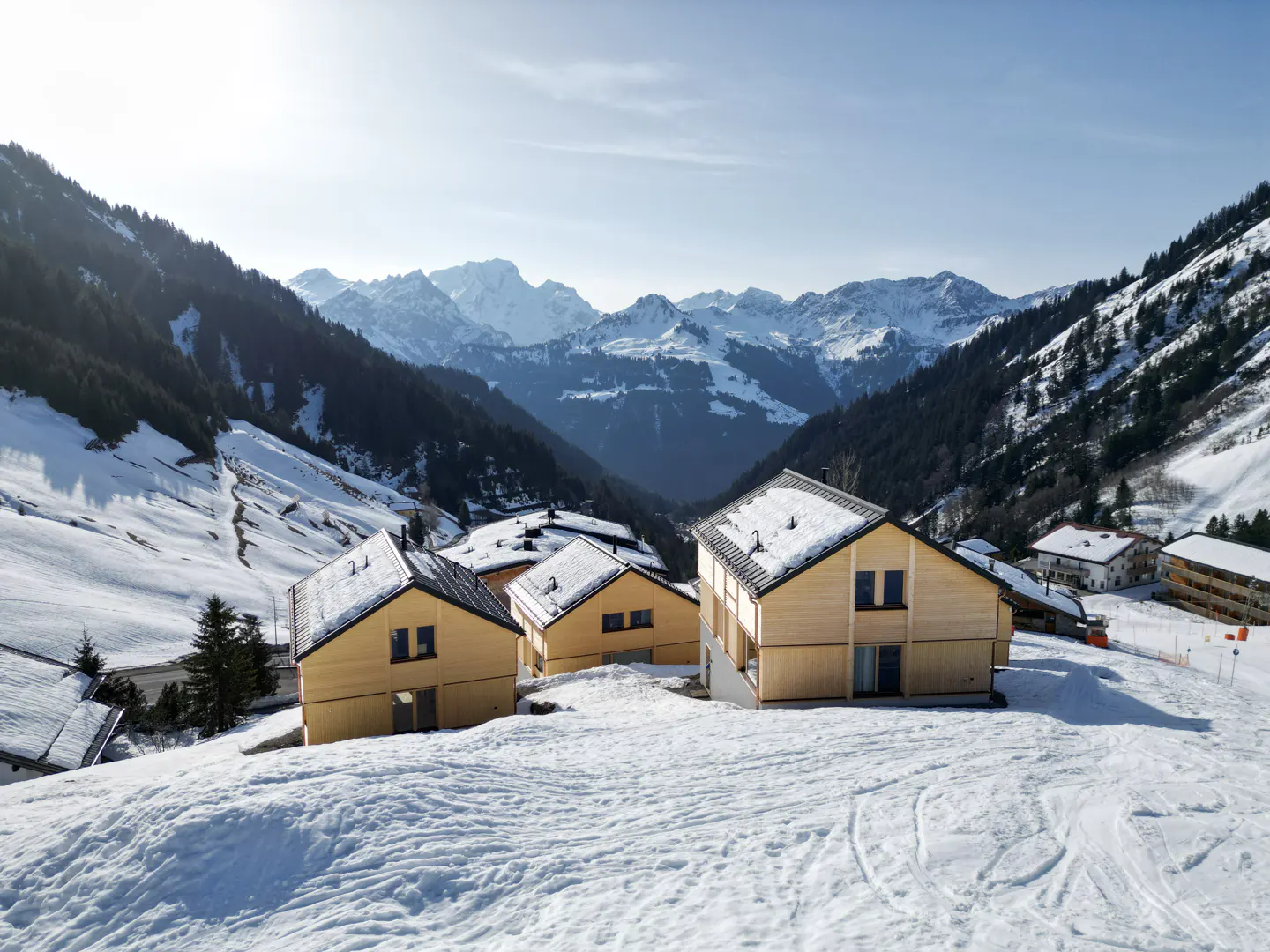 Scenic view of modern wood cabins nestled in a snowy mountain valley, with snow-capped peaks in the background.