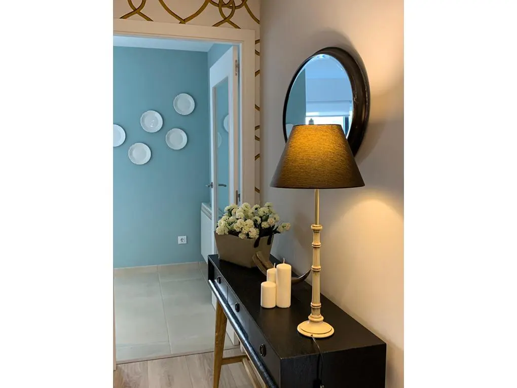 Hallway with a black console table, lamp, mirror, and flowers. A blue room with white plates is visible through the doorway.