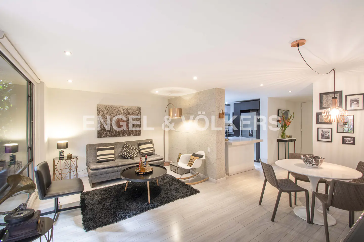 Open-concept living space with gray sofa, black rug, and round table. Dining area with white table and brown chairs. Engel & Völkers logo visible.