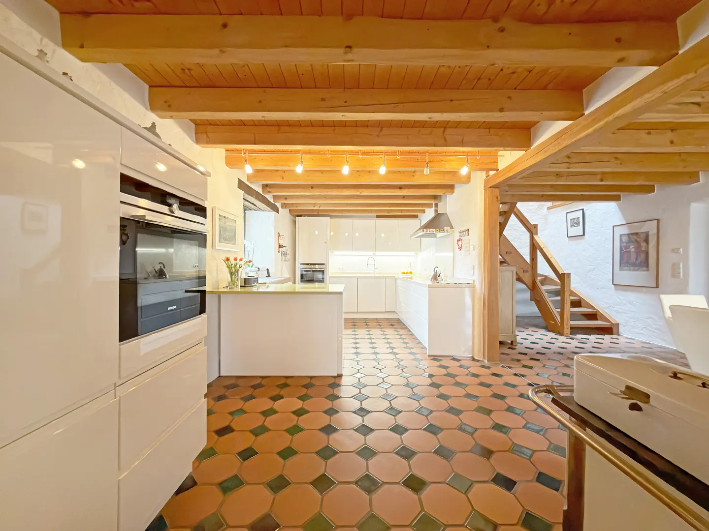A bright kitchen with white cabinets, wood beams, and octagon tile floors. A wooden staircase is visible in the background.