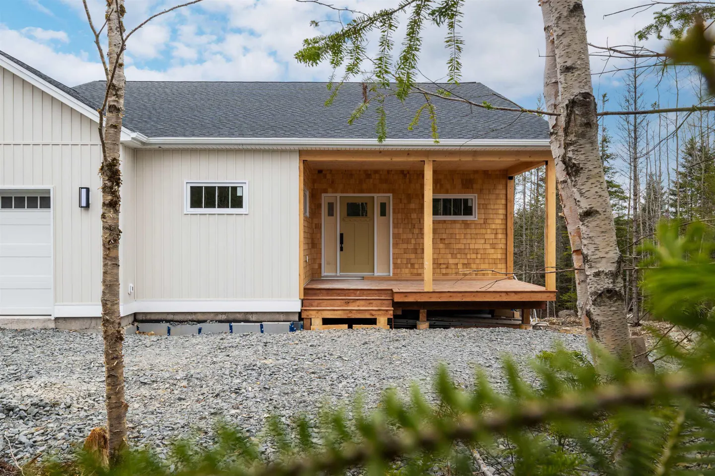 Exterior view of a two-section house with white siding and a cedar shake porch, set on a gravel lot with trees.
