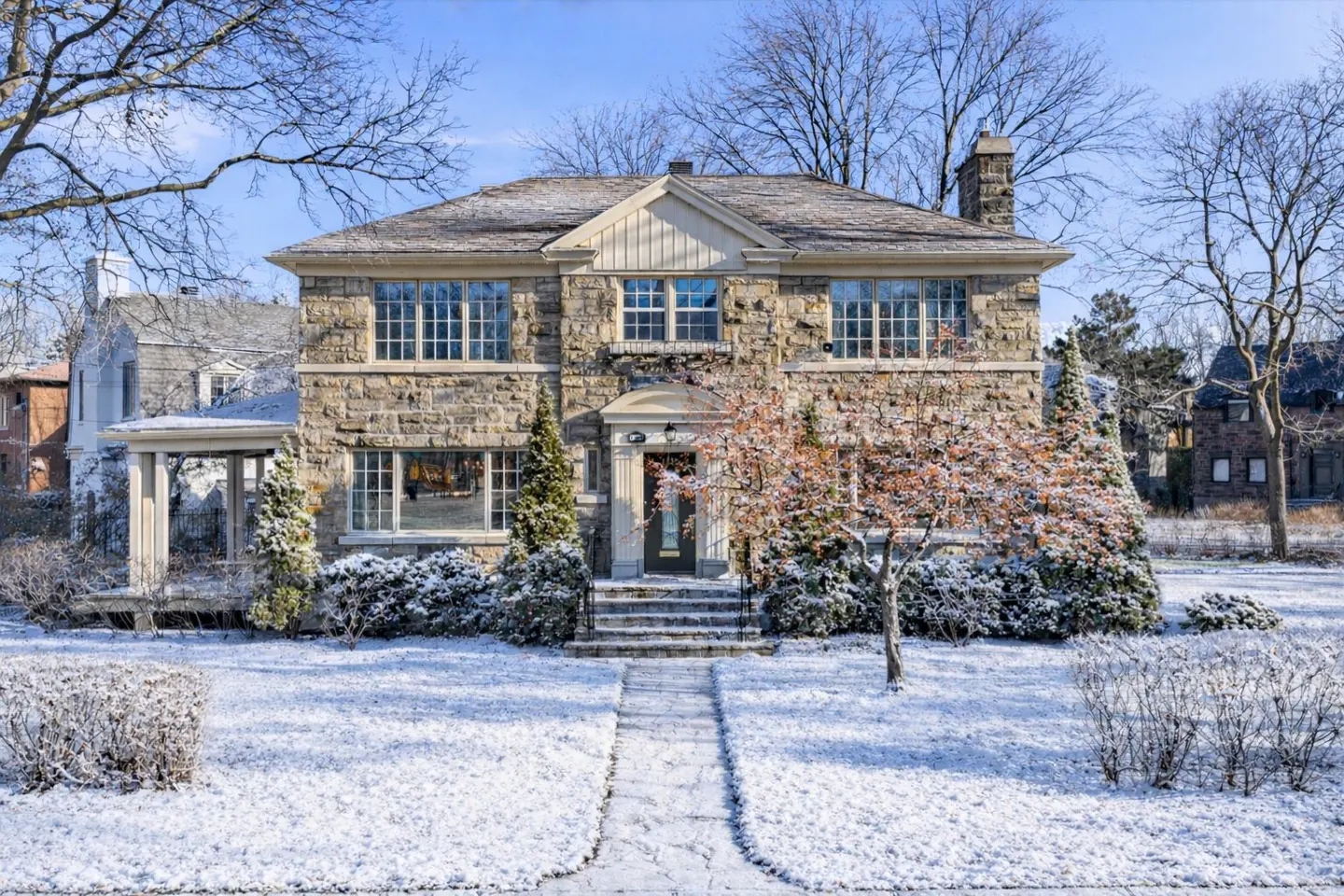 Two-story stone house with a gray roof and a chimney, surrounded by snow-covered trees and bushes. A stone walkway leads to the front door.