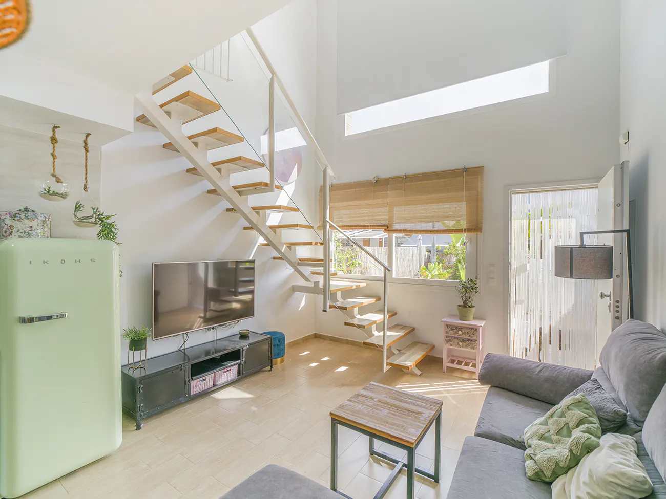 Bright living room with a light green retro fridge, TV, gray sofa, and open staircase with wooden steps.