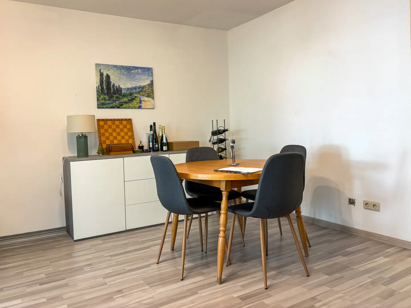 Dining area with a round wooden table, four dark grey chairs, a white sideboard, and a landscape painting on a beige wall.