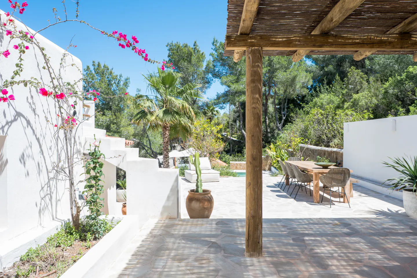 Outdoor patio with a wooden pergola, dining table, and white walls with pink flowers. Lush greenery and a blue sky are visible in the background.