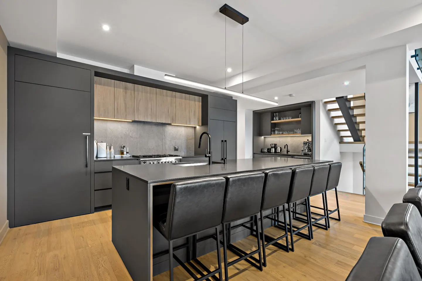 Modern kitchen with gray cabinets, wood accents, and a large island with black leather bar stools. Hardwood floors and a staircase are visible.