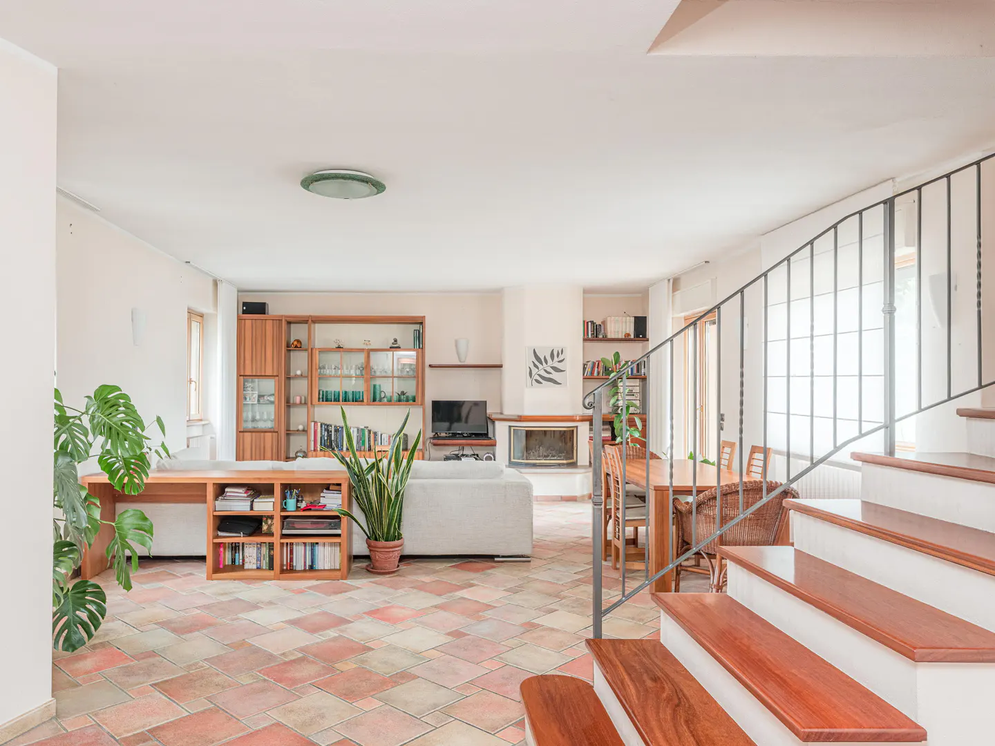 Open-concept living room with terracotta tile floor, white walls, and wooden staircase. A fireplace, bookshelves, and plants add warmth.