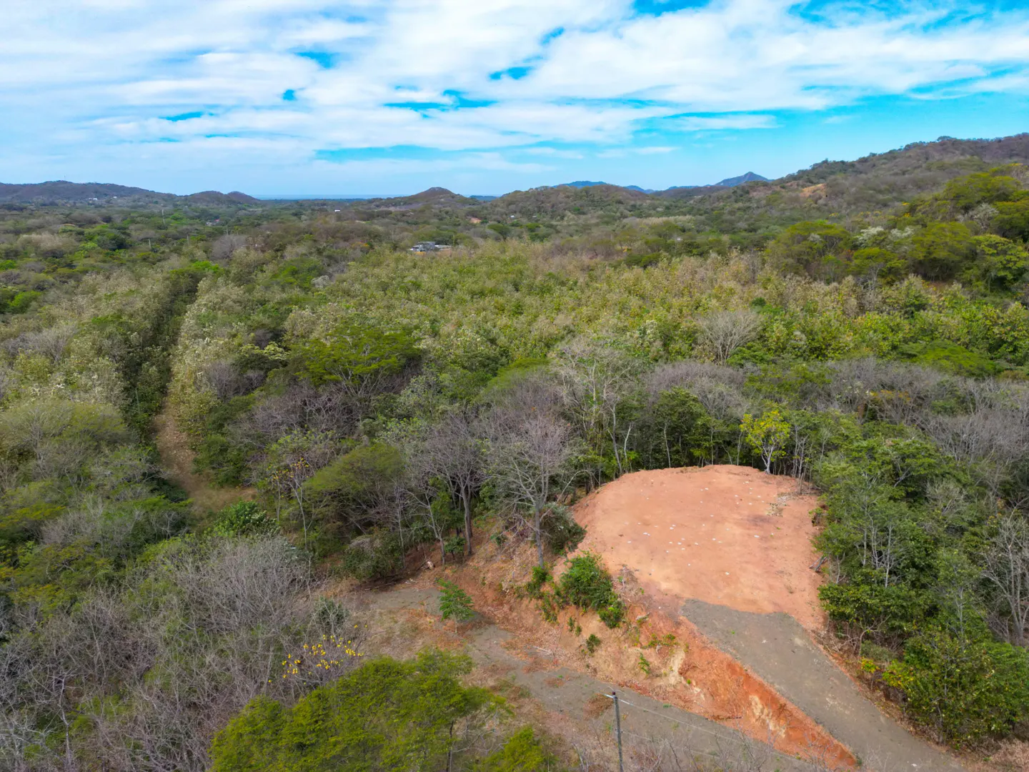 Aerial view of a real estate lot with red soil, surrounded by green trees and mountains under a blue sky.