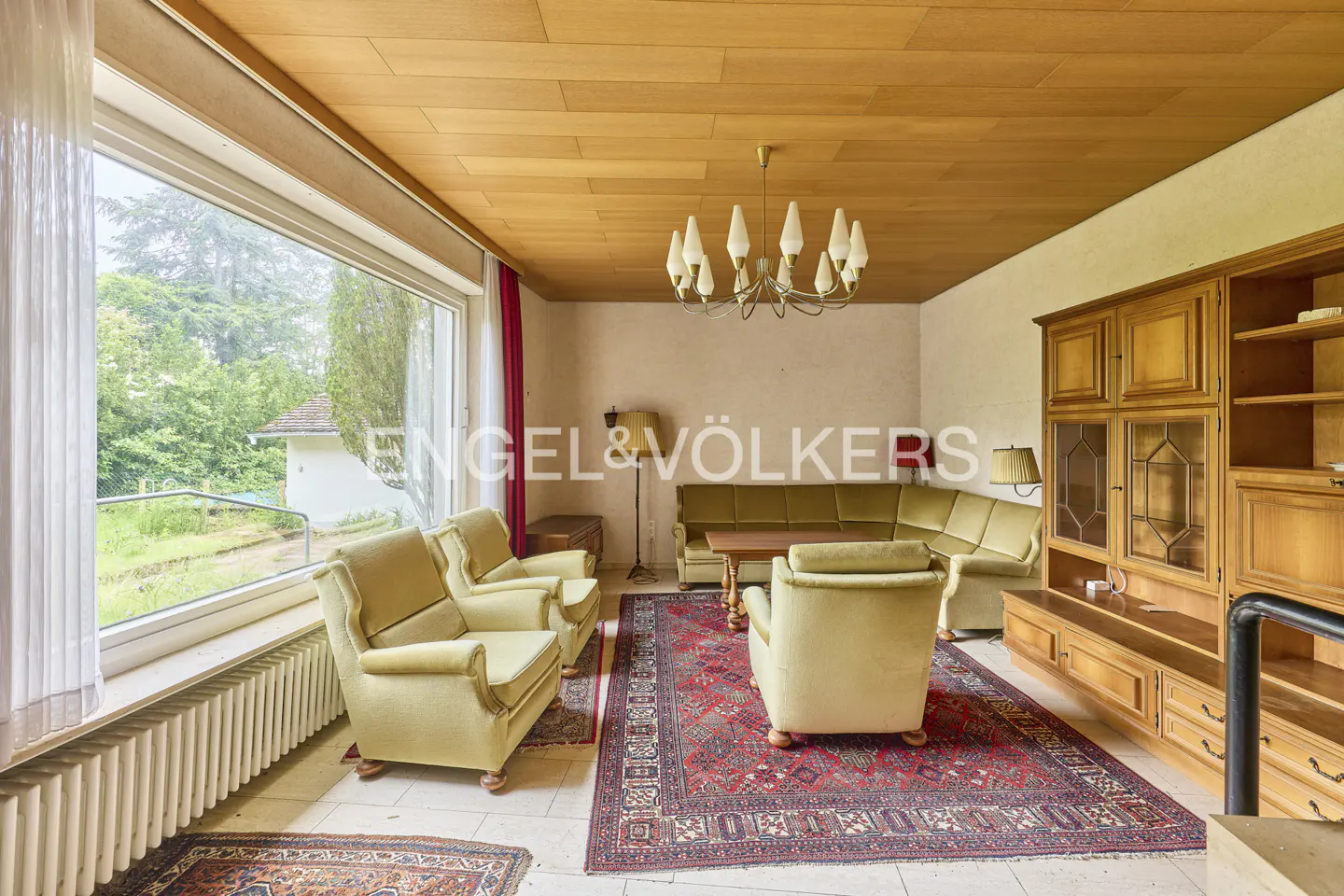 A living room with a wooden ceiling, a large window, a green sofa, and armchairs. A red rug covers the floor.
