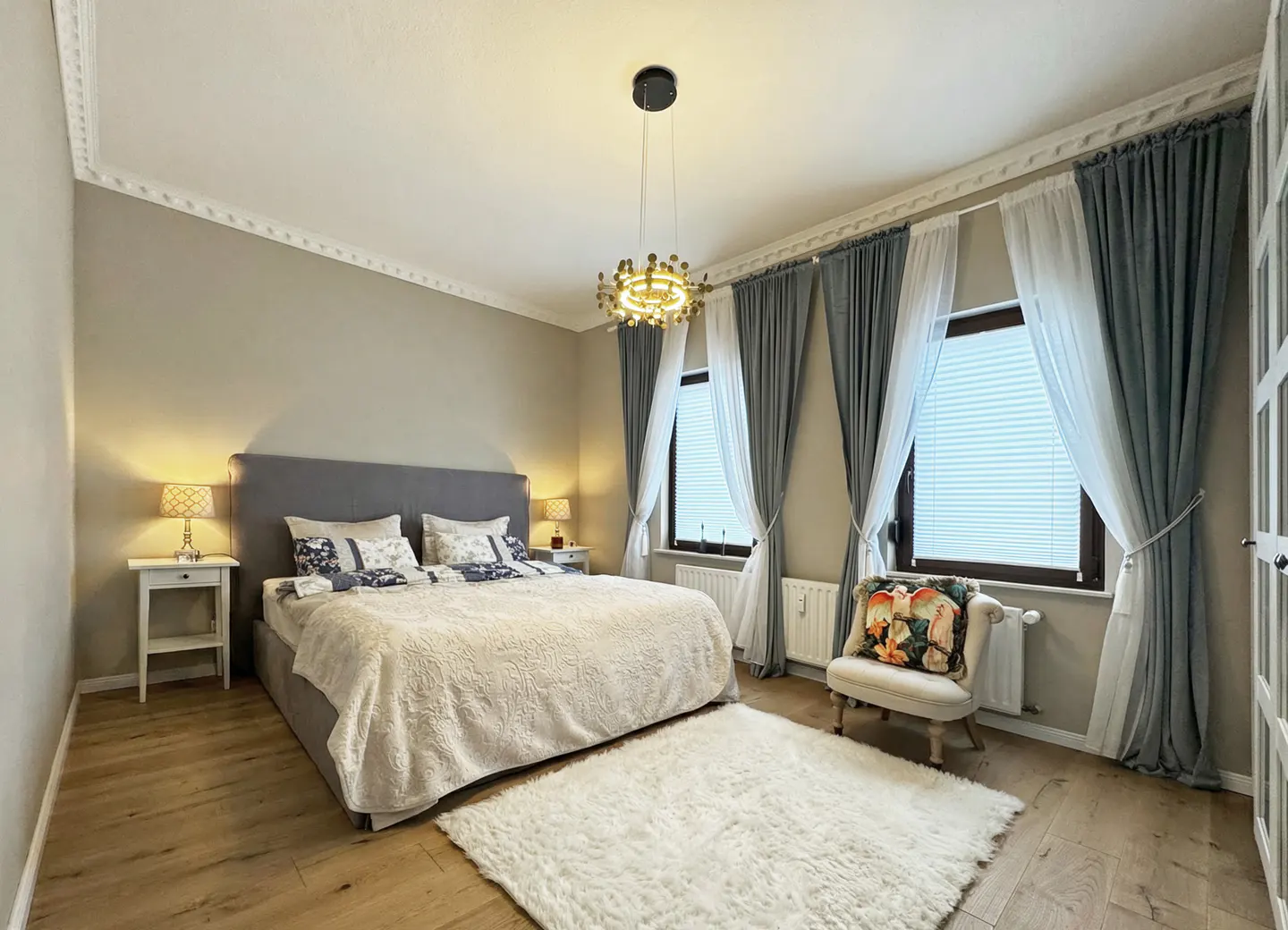 A bedroom with a gray bed, white rug, and gray and white curtains. A gold chandelier hangs from the ceiling.
