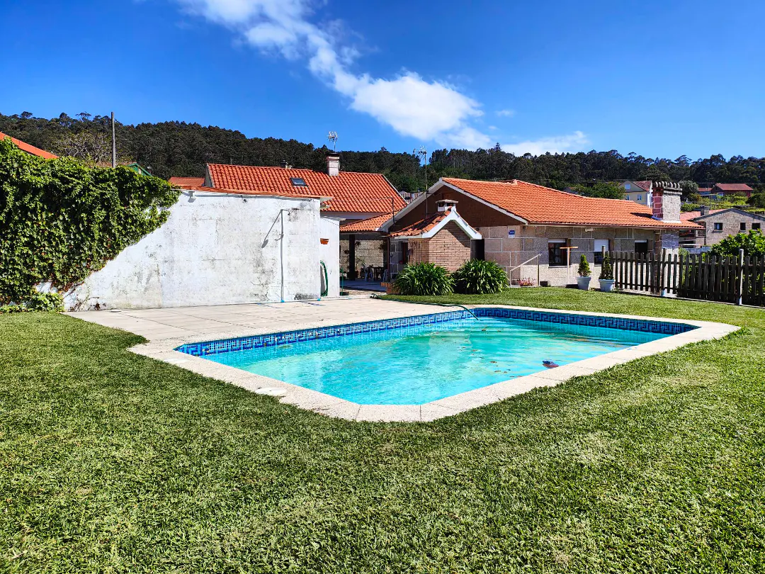 Outdoor pool with blue water, surrounded by green grass and stone patio. Houses with orange tile roofs in the background. Blue sky.