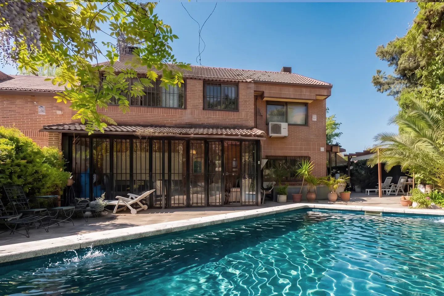 Two-story brick house with a pool in the foreground. The pool is turquoise and reflects the sky. There are trees and plants around the pool.