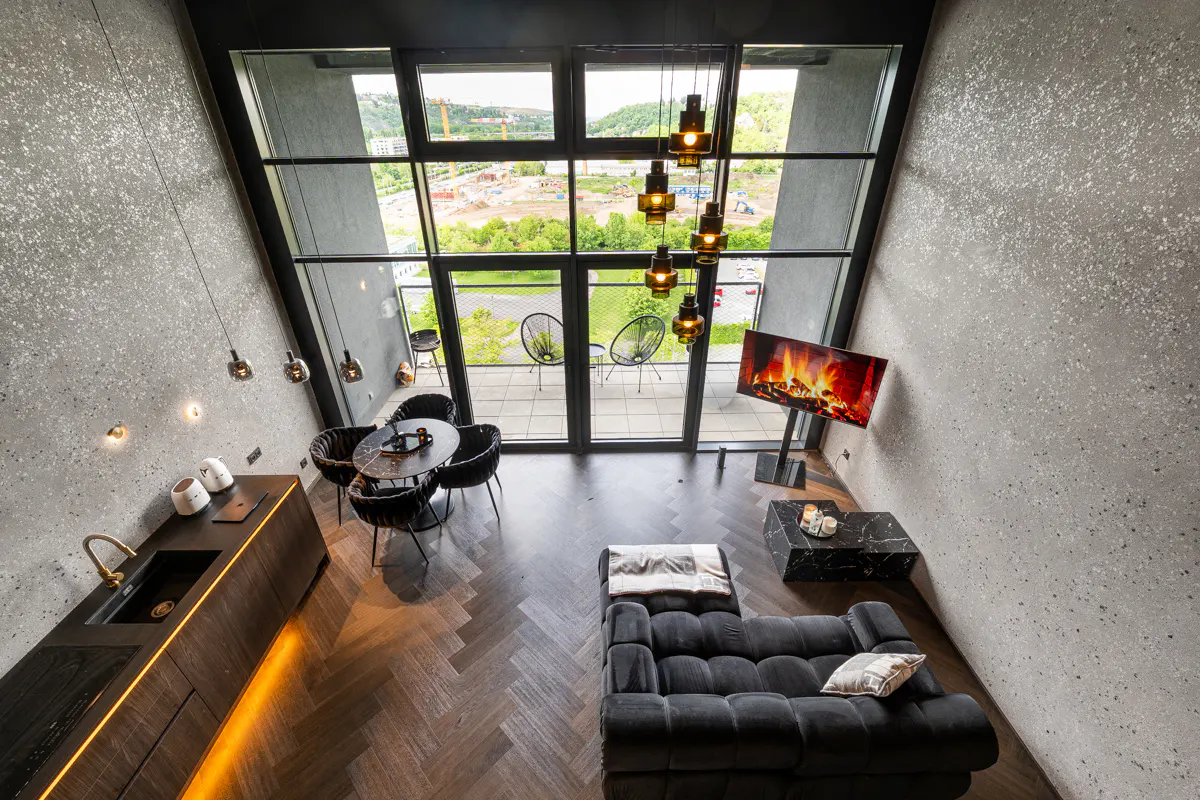 A high-angle view of a modern apartment with a black sofa, kitchen, and a large window overlooking a green landscape.