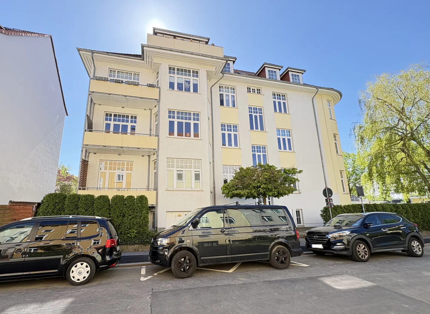 A three-story apartment building with cream and yellow walls, white trim, and three black cars parked in front.