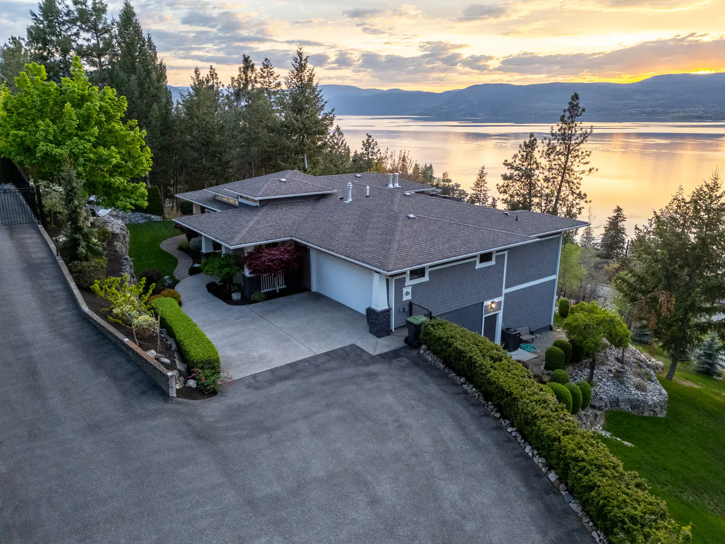 Aerial view of a gray two-story house with a gray roof, a driveway, and a lake in the background at sunset.